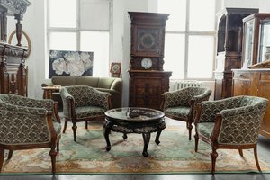 A vintage-style room with four ornate armchairs around a round table on a patterned rug. There's a large wooden cabinet, a painting of flowers, and a green sofa by the window in the background. Natural light filters through multiple windows.