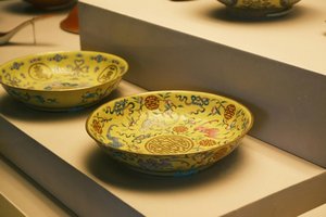 Two ornate yellow ceramic bowls with intricate multicolored designs, including floral and geometric patterns, are displayed on a shelf. The bowls feature motifs of dragons and traditional symbols, showcasing delicate craftsmanship.