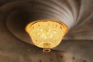 A grand, ornate crystal chandelier with warm lighting is mounted on a decorative ceiling with a swirling pattern.