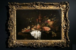 A still life painting featuring a lavish spread of food, including meats, fruits, and vegetables, set on a table with a white cloth. The scene is framed in an ornate, gilded frame against a dark background.