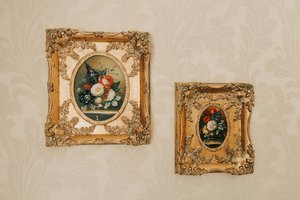 Two ornate gold-framed paintings of flowers hang on a patterned wallpapered wall. The left painting is larger, featuring a bouquet of roses and other flowers in a vase; the right painting is smaller with a similar floral arrangement.