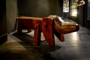 A wooden sculpture of a dog on a dark floor, resembling a bench. The sculpture features a simplistic, artistic design with a long body, four legs, and a tail. The background includes a dimly lit hallway with display cases.