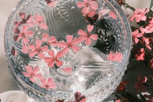 A decorative glass bowl filled with water, adorned with floating pink flowers. Surrounded by more pink flowers on a soft, light-colored fabric background, creating an elegant and serene atmosphere.